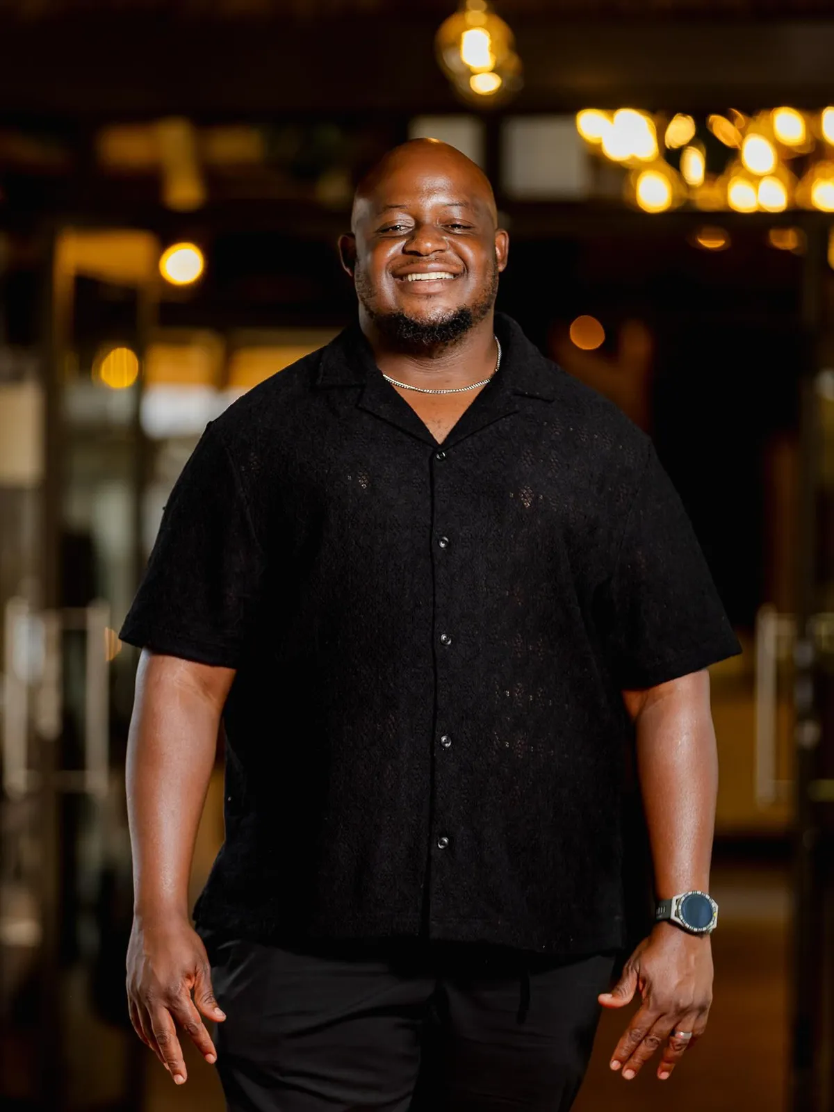 Oscar Omuhle smiling in a black shirt, warm lights behind him